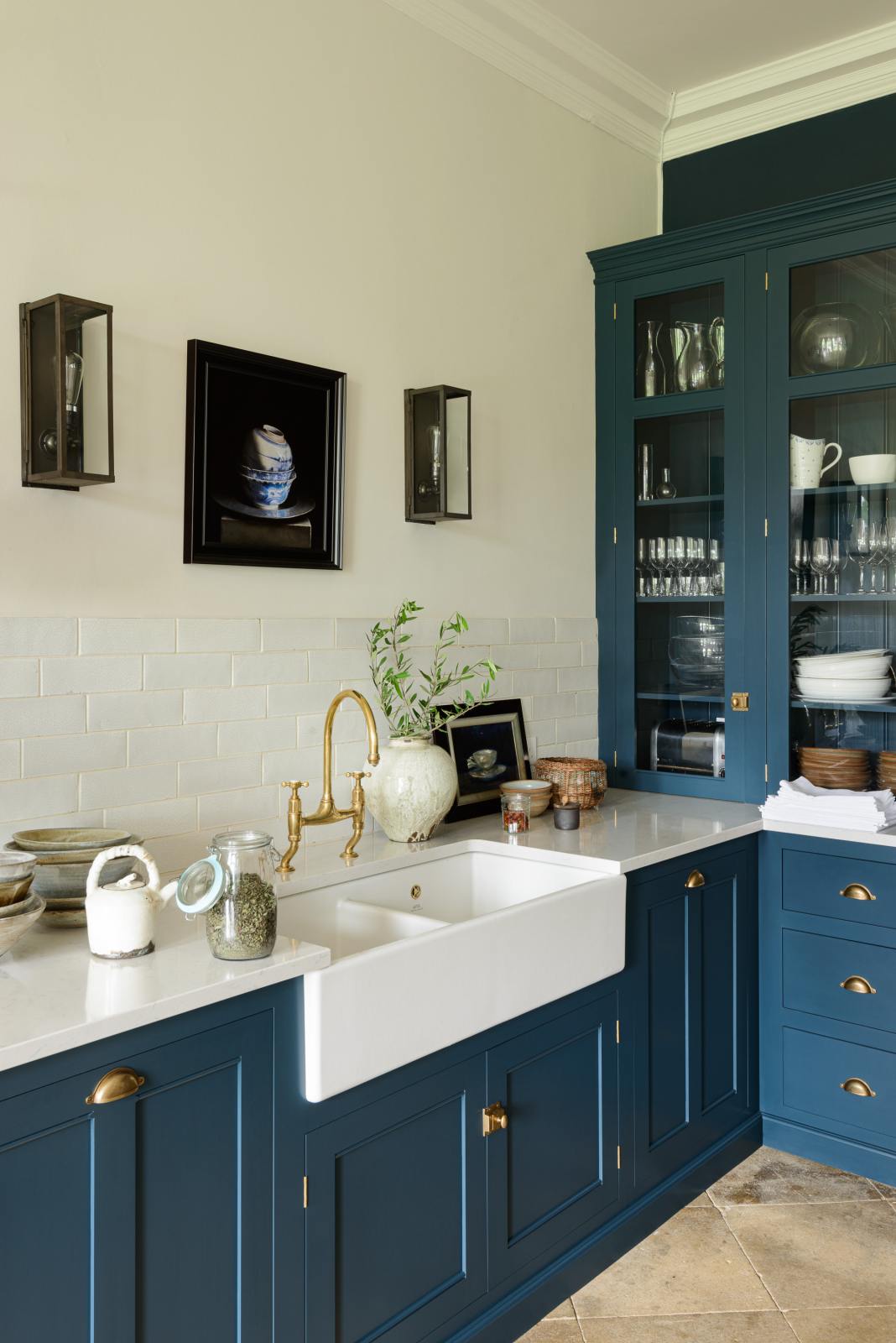 An oblique view of a classic English, Bond Street blue kitchen features a cooker and sink run with a Silestone worktop, glazed counter cabinets, a double sink, and an aged brass Ionian tap. The renovated medieval house is elegantly adorned with modern still-life paintings, artisan ceramics, and vibrant green plants, seamlessly blending the historical with the contemporary.