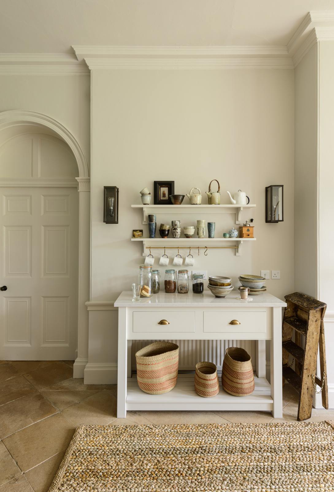 A small section of the renovated medieval house’s kitchen is a drinking station with a white linen prep table, a Silestone worktop, and shelves with a brass hanging rail. It is elegantly adorned with vintage furniture, artisan ceramics, mugs, and a collection of teas and coffee beans, seamlessly blending the historical with the contemporary.
