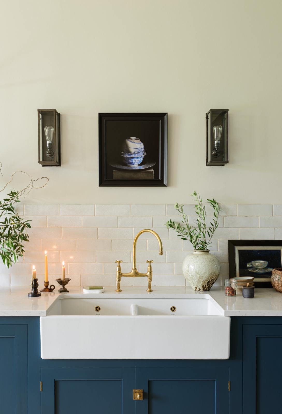 A closer look at a classic English, Bond Street blue kitchen features a cooker and sink run with a Silestone worktop, a double sink, an aged brass Ionian tap, and white crackle metro splashback tiles. The renovated medieval house is elegantly adorned with modern still-life paintings, artisan ceramics, and vibrant green plants, seamlessly blending the historical with the contemporary.