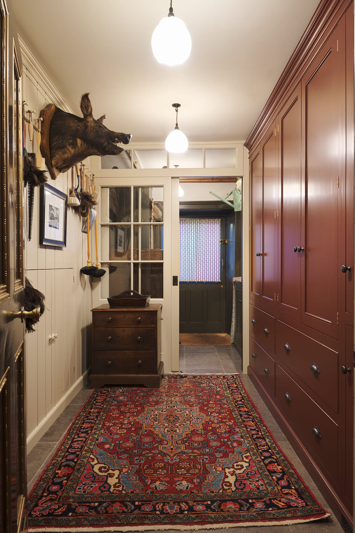 A practical Refectory red corridor with a classic carcass with a double door, full-height cupboards, and peg rail. Pridefully displaying a taxidermy boar, a large rug, and collectable antiques, it embraces a unique museum-like social space.