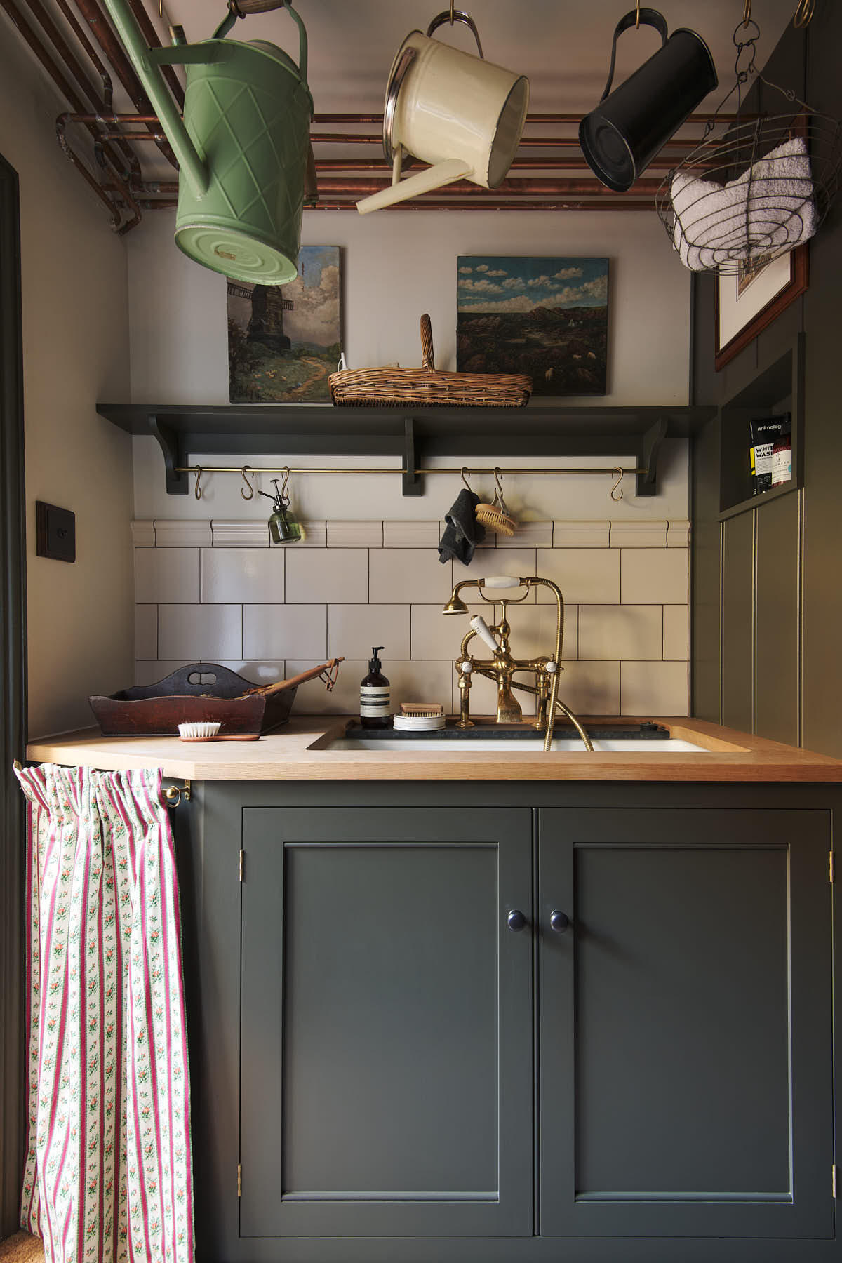A beautifully cosy and practical Bakehouse green scullery with an undermount sink cabinet, shelf with brass rails, and a cream subway splashback. Lovingly adorned with antique watering cans, paintings, vintage items, it creates a functional little nook.