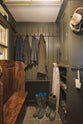 A beautifully cosy and practical Bakehouse green boot room with an open cabinet with curtain railing, boiler storage, and a peg rail. Lovingly adorned with an antique pew, muddy boots, and different size boots, it creates a functional little nook.