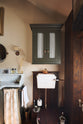 A glamorous Bakehouse green bathroom with a bespoke sink cabinet with a marble worktop, shelf, splashback, and a wall cabinet with ruched curtains. Beautifully detailed cream wallpaper, a simple mirror, an elegant painting, and a touch of greenery from a houseplant come together to create a gorgeously chic space.