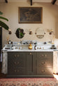 A glamorous Bakehouse green bathroom with a bespoke sink cabinet with a marble worktop, shelf and splashback. Beautifully detailed cream wallpaper, a simple mirror, an elegant painting, and a touch of greenery from a houseplant come together to create a gorgeously chic space.