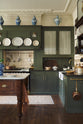 A quiet corner of an eccentric yet grand Bakehouse Green Classic English Kitchen features an L-shaped sink and cooker run with black granite worktop, a glazed wall cupboard, an overmantle with cream subway splashback tiles, and an Heirloom dairy table. Pridefully displaying taxidermy, artisan ceramics, and collectable antiques, it embraces a unique museum-like social kitchen space.