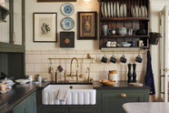 A closer look at the eccentric yet grand Bakehouse Green Classic English Kitchen features an L-shaped sink and cooker run with black granite worktop, a glazed wall cupboard, and an aged brass Ionian tap. Pridefully displaying taxidermy, artisan ceramics, and collectable antiques, it embraces a unique museum-like social kitchen space.