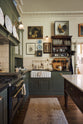 An eccentric yet grand Bakehouse Green Classic English Kitchen features an L-shaped sink and cooker run with black granite worktop, a glazed wall cupboard, an overmantle with cream subway splashback tiles, and an Heirloom dairy table. Pridefully displaying taxidermy, artisan ceramics, and collectable antiques, it embraces a unique museum-like social kitchen space.