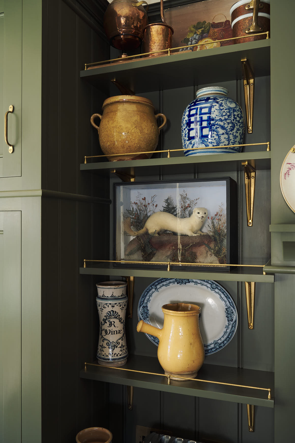 A closer look at the eccentric yet grand Bakehouse Green Classic English Kitchen’s shelves with the classic shelf brackets and aged brass railing. Pridefully displaying taxidermy, artisan ceramics, and collectable antiques, it embraces a unique museum-like social kitchen space.