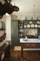 A quiet corner of an eccentric yet grand Bakehouse Green Classic English Kitchen features an L-shaped sink and cooker run with black granite worktop, a full-height cabinet, an overmantle with cream subway splashback tiles, and an Heirloom dairy table. Pridefully displaying taxidermy, artisan ceramics, and collectable antiques, it embraces a unique museum-like social kitchen space.