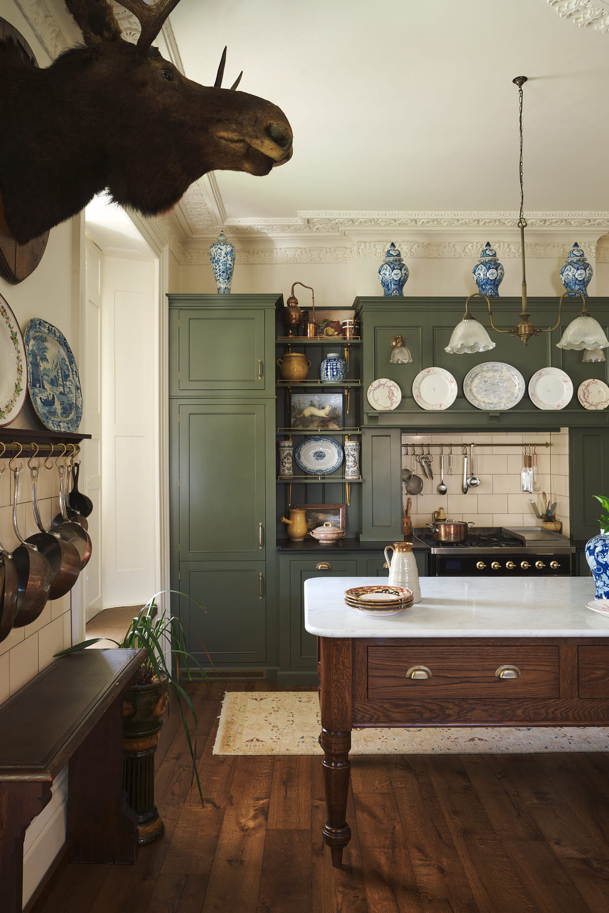 A quiet corner of an eccentric yet grand Bakehouse Green Classic English Kitchen features an L-shaped sink and cooker run with black granite worktop, a full-height cabinet, an overmantle with cream subway splashback tiles, and an Heirloom dairy table. Pridefully displaying taxidermy, artisan ceramics, and collectable antiques, it embraces a unique museum-like social kitchen space.