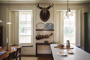 An eccentric yet grand Bakehouse Green Classic English Kitchen features an oiled oak shelf with brass hanging rail, and an Heirloom dairy table. Pridefully displaying a taxidermy moose, antique furniture, and artisan ceramics, it embraces a unique museum-like social kitchen space.
