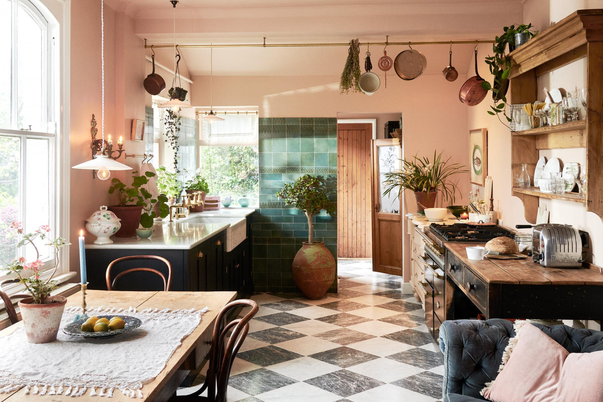 A bright peach-coloured room with a chequerboard marble floor and an eclectic collection of vintage furniture, including a table and chairs, a prep table and a decorative set of drawers. In the left corner is a modest sink run with black painted Classic English cupboards, a Carrara marble worktop and brass fittings. Pots and pans hang from a brass ceiling rail, mixed with a collection of vintage milk glass pendant lights.
