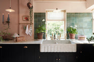 A closer look at the bright peach-coloured kitchen reveals a Carrara marble worktop with a double fluted sink and twin aged brass Mayan taps. Vintage teal lace-market splashback tiles add character, paired with a small brass rail holding a teal teacup and tea towels. The space is styled with an eclectic mix of vintage pieces, including a wall-mounted candle holder, a radio, a gold-framed painting of a tomato, and potted plants dotted across the counter.