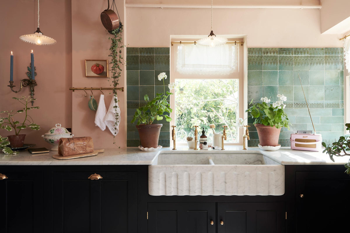 A closer look at the bright peach-coloured kitchen reveals a Carrara marble worktop with a double fluted sink and twin aged brass Mayan taps. Vintage teal lace-market splashback tiles add character, paired with a small brass rail holding a teal teacup and tea towels. The space is styled with an eclectic mix of vintage pieces, including a wall-mounted candle holder, a radio, a gold-framed painting of a tomato, and potted plants dotted across the counter.