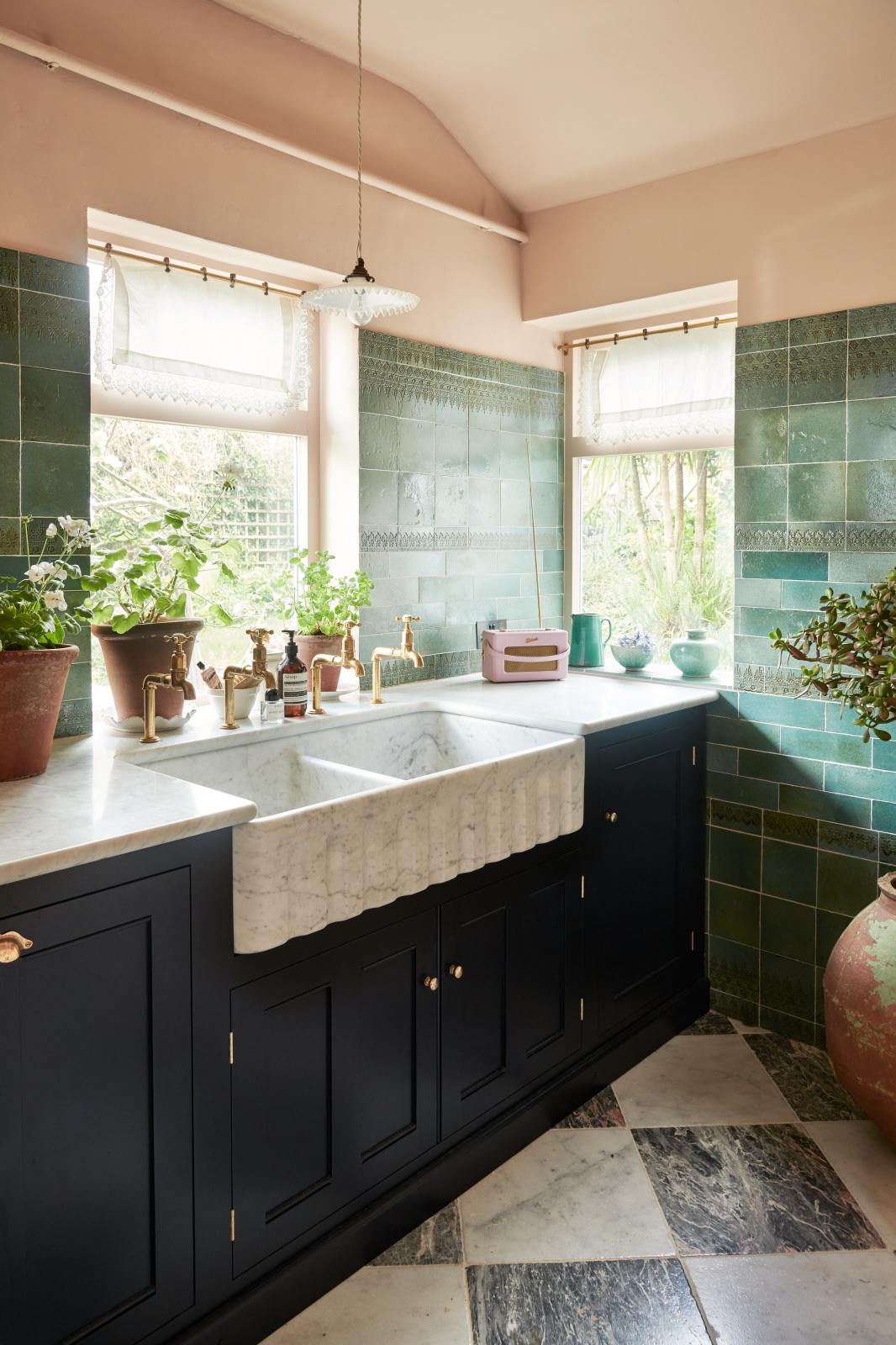 An angled view of the bright peach-coloured kitchen reveals a Carrara marble worktop with a double fluted sink and twin aged brass Mayan taps. Vintage teal lace-market splashback tiles add character. The space is styled with an eclectic mix of vintage pieces, including a radio, potted plants scattered across the counter, and a large one on the floor.