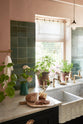 An angled view of the bright peach-coloured kitchen reveals a Carrara marble worktop with a double fluted sink and twin aged brass Mayan taps. Vintage teal lace-market splashback tiles add character, paired with a small brass rail holding a teal teacup and tea towels. The space is styled with a ceramic jug holding blossomed branches, potted plants scattered across the counter, and a charcuterie board with sourdough bread and a jar of orange marmalade.