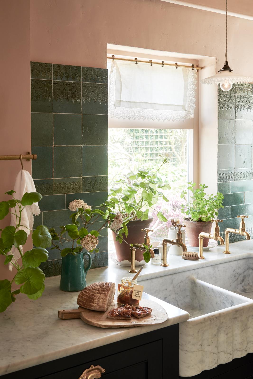 An angled view of the bright peach-coloured kitchen reveals a Carrara marble worktop with a double fluted sink and twin aged brass Mayan taps. Vintage teal lace-market splashback tiles add character, paired with a small brass rail holding a teal teacup and tea towels. The space is styled with a ceramic jug holding blossomed branches, potted plants scattered across the counter, and a charcuterie board with sourdough bread and a jar of orange marmalade.