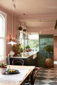 A closer look at the bright peach-coloured kitchen reveals a Carrara marble worktop with a double fluted sink and twin aged brass Mayan taps. Vintage teal lace-market splashback tiles add character, paired with a small brass rail holding a teal teacup and tea towels. The space is styled with an eclectic mix of vintage pieces, including a dining table, a pink radio, Bentle wood chairs, and potted plants dotted across the counter, with a large one on the floor.