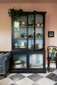 A closer look at the painter’s black curiosity cupboard reveals a Trinity Blue interior that beautifully highlights a collection of quirky ceramic plates, bowls, and mugs. The bright peach-coloured room features a chequerboard marble floor and an eclectic collection of vintage furniture, including a velvet blue sofa, a small cupcake painting, blossoming branches in a teal jug, and a wooden chair.