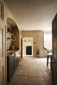 Looking through the entranceway to a rustic basement kitchen with sandy-coloured walls and ceiling, chipped paintwork and a worn stone floor. On the left is a short run of elegant black cupboards with a soft sheen and a mottled copper worktop. Ahead, light pours into the room through a large window with original wavy Georgian panes and an open doorway leading outside to a stone courtyard.