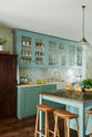An angled view of a spacious yet modest Trinity Blue classic English kitchen reveals a Silestone worktop sink run with a single fluted Belfast sink and aged brass Ionian tap, glazed cupboards, a copper prep table, and deVOL wooden bum stools—all finished with a white porcelain splashback. Beautifully adorned with wild white flowers in a vase, glass pendant lights, a watermelon, papaya, live basil, dried ingredients in glass jars, the room feels cosy, warm, and inviting.