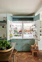 A spacious yet modest Trinity Blue classic English kitchen features a Silestone worktop cooker run, an overmantle canopy with a white porcelain splashback, an open large full-height cabinet housing a fridge/freezer, a copper prep table, and deVOL wooden bum stools. Lovingly decorated with a watermelon, live basil, and dried ingredients in glass jars, the room feels humble, warm, and inviting.