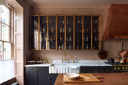 A bold and dramatic Haberdasher’s kitchen with a sink and cooker run, an Arabescato worktop and single fluted sink, an aged brass Ionian tap, a glazed wall cupboard, a large copper canopy, and a kitchen island with a copper worktop. Adorned with copper pans, artisan ceramics, labelled glass jars, and a quirky framed illustration, creating a local bristo bar in a Manhattan space.