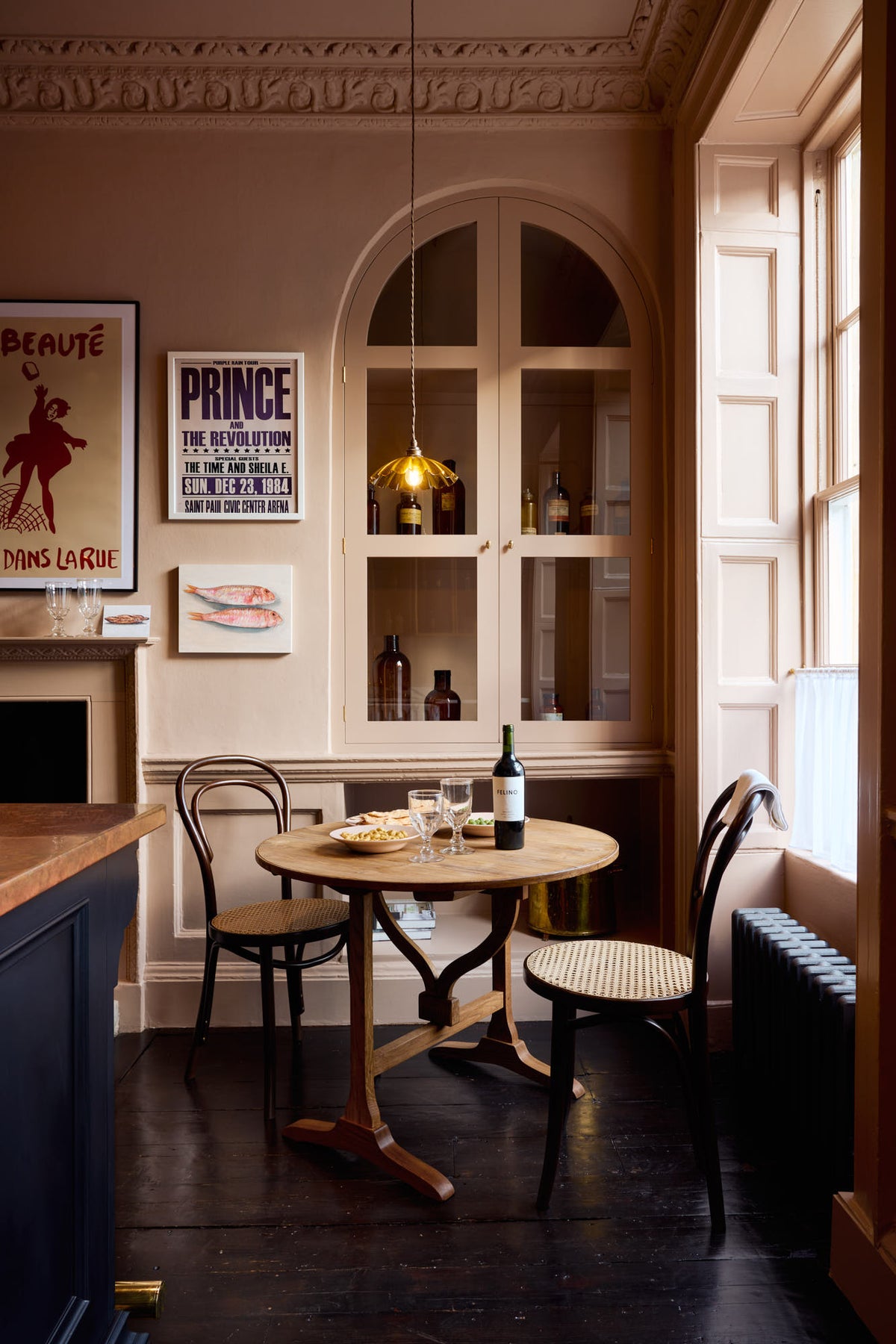 A quiet corner of a bold and dramatic Haberdasher’s kitchen features a deVOL petite vineyard table, Bentwood café chairs, and a petite frilly chartreuse light. Adorned with a delicious bottle of wine, drinking glasses, hors d’oeuvres, quirky framed illustrations and posters, and labelled glass jars, this space creates a local bistro bar atmosphere in Manhattan.