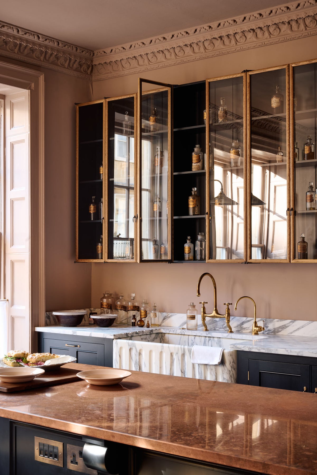 An oblique view of a bold and dramatic Haberdasher’s kitchen with a sink and cooker run, an Arabescato worktop and single fluted sink, an aged brass Ionian tap, a glazed wall cupboard, and a kitchen island with a copper worktop. Adorned with artisan ceramics, labelled glass jars, and a colourful sandwich, creating a local bristo bar in a Manhattan space.