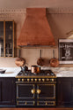 A close-up of a bold and dramatic Haberdasher’s kitchen with a sink and cooker run, an Arabescato worktop, a large copper canopy, aged brass hanging rails, and a black with golden accent gas cooker. Adorned with copper pans, artisan ceramics, labelled glass jars, a milk glass task light, and a quirky framed illustration, creating a local bristo bar in a Manhattan space.