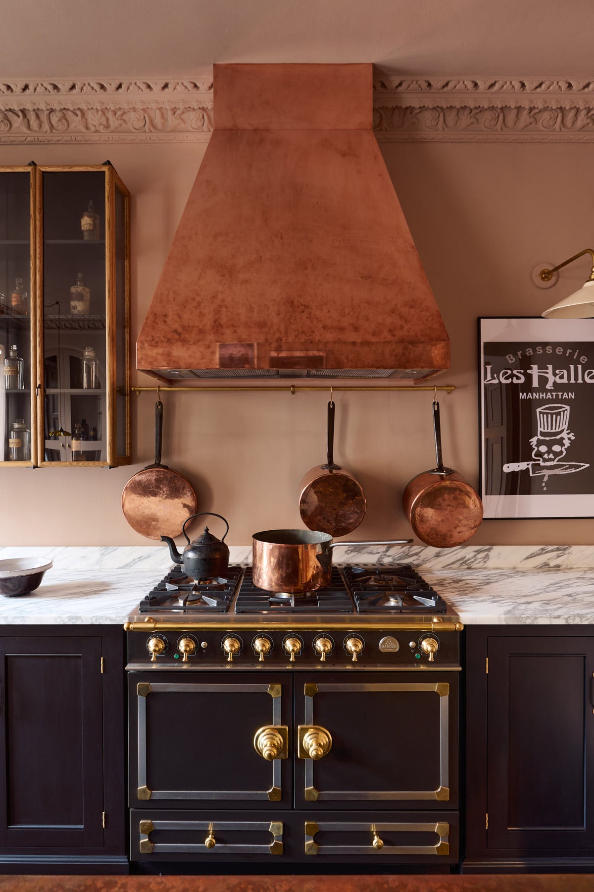 A close-up of a bold and dramatic Haberdasher’s kitchen with a sink and cooker run, an Arabescato worktop, a large copper canopy, aged brass hanging rails, and a black with golden accent gas cooker. Adorned with copper pans, artisan ceramics, labelled glass jars, a milk glass task light, and a quirky framed illustration, creating a local bristo bar in a Manhattan space.