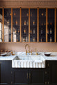 A closer look at a bold and dramatic Haberdasher’s kitchen with a sink and cooker run, an Arabescato worktop and single fluted sink, an aged brass Ionian tap, and a glazed wall cupboard. Adorned with artisan ceramics, labelled glass jars, and a quirky framed illustration, creating a local bristo bar in a Manhattan space.