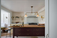 A warm, inviting, muted yellow classic English kitchen features an L-shaped cooker and sink run with a Carrara marble worktop, a large freestanding maroon prep table with a solid oak worktop and a breakfast nook. Adorned with deVOL dark creedy stools, cherished muted ceramic plates, bowls, a deVOL brass heirloom gaselier light, freshly baked baguettes, wine, and a charcuterie board, this kitchen feels humble and inviting for a family that enjoys cooking.