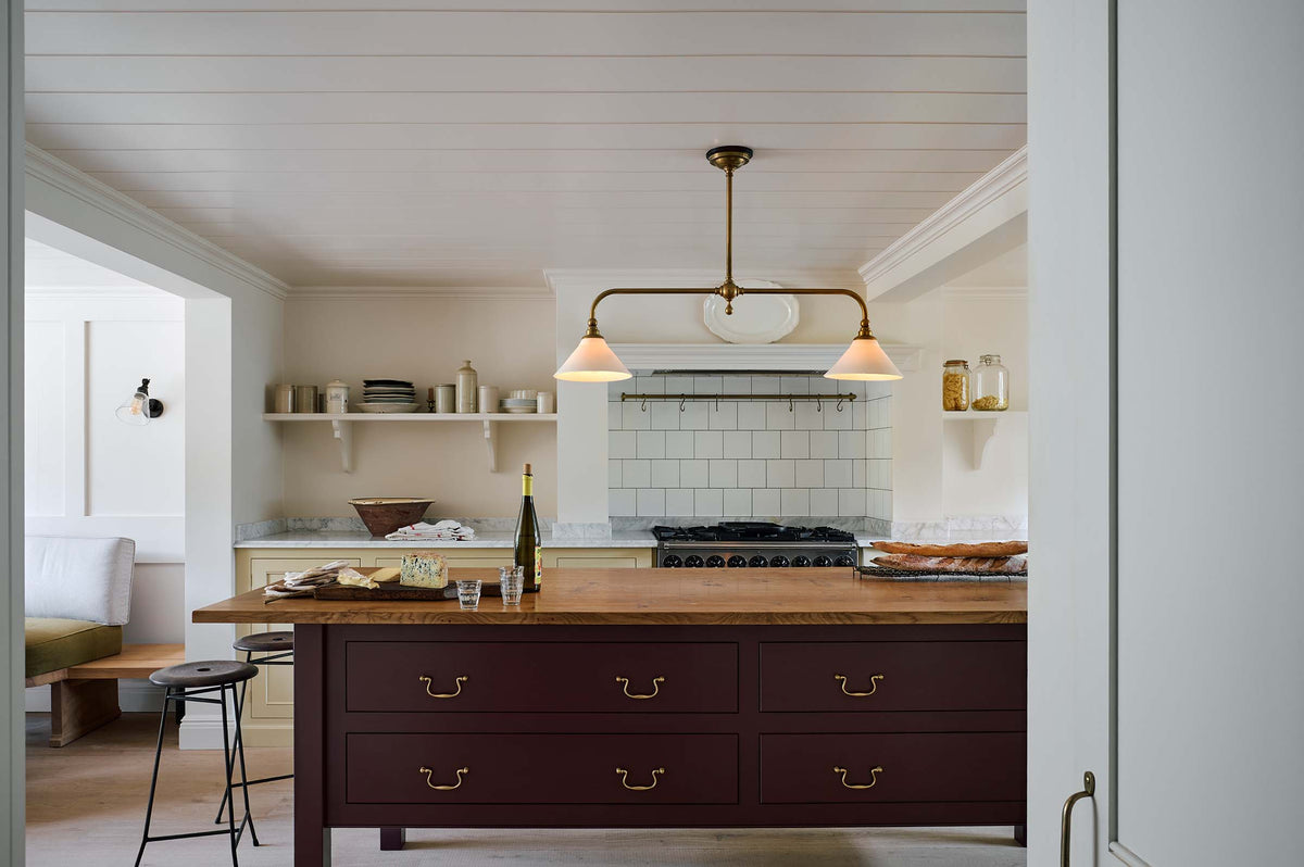A warm, inviting, muted yellow classic English kitchen features an L-shaped cooker and sink run with a Carrara marble worktop, a large freestanding maroon prep table with a solid oak worktop and a breakfast nook. Adorned with deVOL dark creedy stools, cherished muted ceramic plates, bowls, a deVOL brass heirloom gaselier light, freshly baked baguettes, wine, and a charcuterie board, this kitchen feels humble and inviting for a family that enjoys cooking.