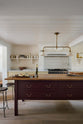 A warm, inviting, muted yellow classic English kitchen features an L-shaped cooker and sink run with a Carrara marble worktop, a large freestanding maroon prep table with a solid oak worktop and a breakfast nook. Adorned with deVOL dark creedy stools, cherished muted ceramic plates, bowls, a deVOL brass heirloom gaselier light, freshly baked baguettes, wine, and a charcuterie board, this kitchen feels humble and inviting for a family that enjoys cooking.