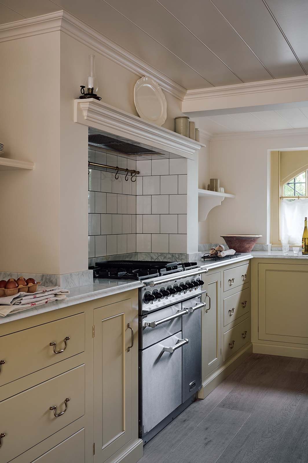 A close and oblique view of a warm, inviting, muted yellow classic English kitchen features an L-shaped cooker and sink run with a Carrara marble worktop, overmantle canopy with white porcelain splashback tiles, and a silver gas cooker. Adorned with cherished, muted ceramic plates, bowls, eggs, freshly baked baguettes, wine, and deVOL aged brass hanging rails, this kitchen feels humble and inviting for a family that enjoys cooking.