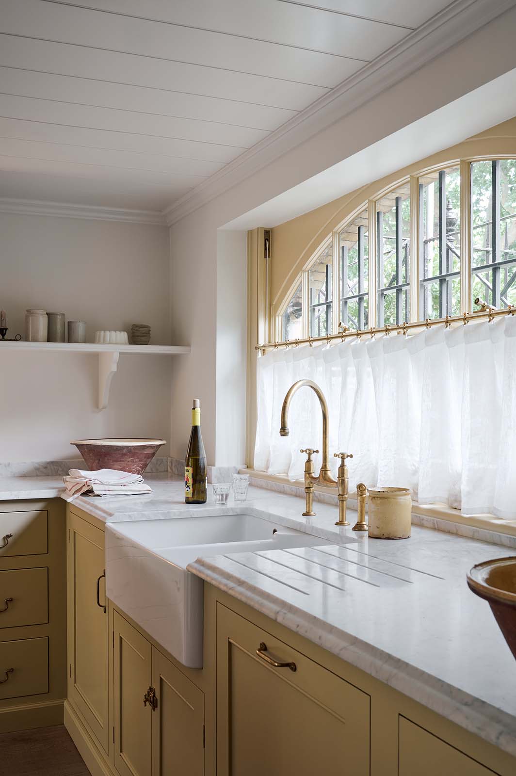 A close and oblique view of a warm, inviting, muted yellow classic English kitchen features an L-shaped cooker and sink run with a Carrara marble worktop and a large freestanding maroon prep table with a solid oak worktop. The double Belfast sink with an aged brass Ionian tap sits in front of a semi-circular window with an aged brass curtain rail at the bottom half, allowing privacy and ample natural light to brighten the room.