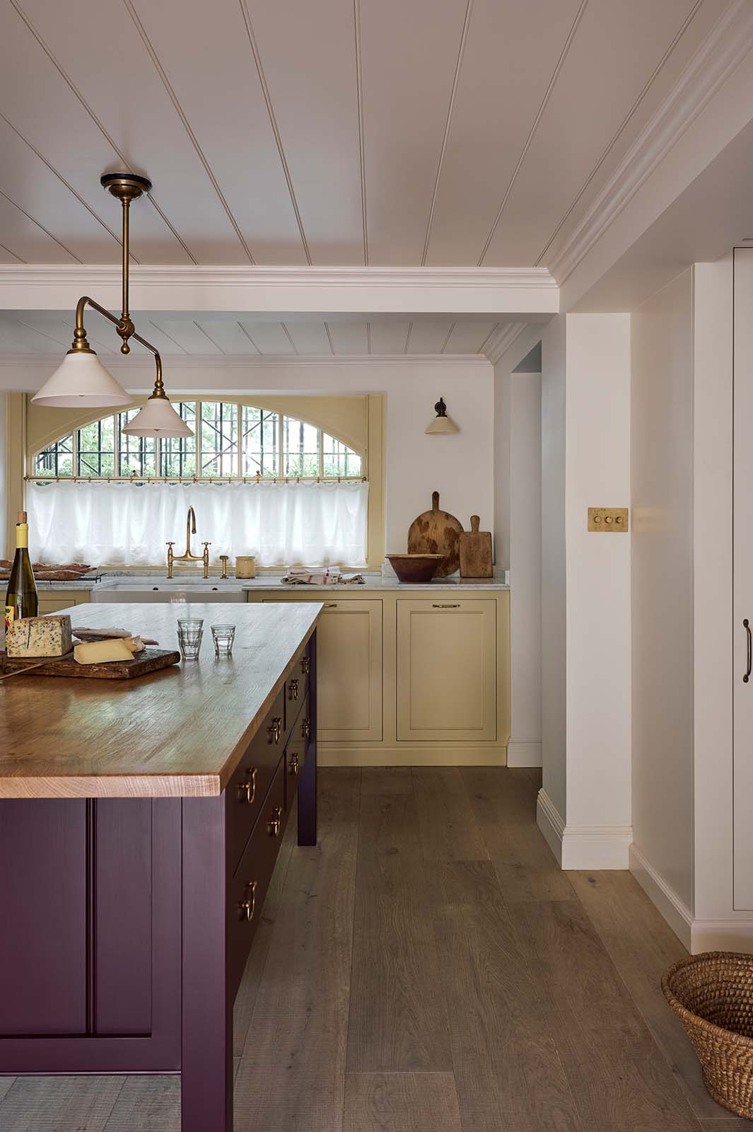 A warm, inviting, muted yellow classic English kitchen features an L-shaped cooker and sink run with a Carrara marble worktop, a large freestanding maroon prep table with a solid oak worktop and a breakfast nook. Adorned with a cherished muted ceramic bowl, a deVOL brass heirloom gaselier light, freshly baked baguettes, wine, and a charcuterie board, this kitchen feels humble and inviting for a family that enjoys cooking.