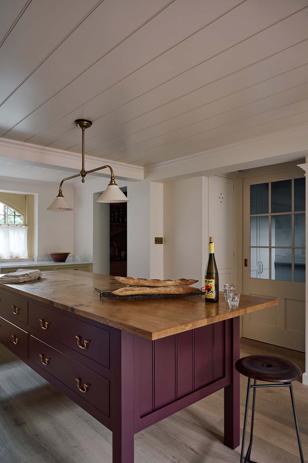 An oblique view of a warm, inviting, muted yellow classic English kitchen features an L-shaped cooker and sink run with a Carrara marble worktop, a large freestanding maroon prep table with a solid oak worktop and a breakfast nook. Adorned with deVOL dark creedy stool, a cherished muted bowl, a deVOL brass heirloom gaselier light, freshly baked baguettes, and wine, this kitchen feels humble and inviting for a family that enjoys cooking.