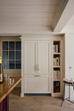 In a quiet corner of a warm, inviting, muted classic English kitchen features a large freestanding maroon prep table with a solid oak worktop and a breakfast nook, a full-height fridge and freezer cupboard and tray slots. Adorned with a deVOL dark creedy stool, books, woods chopping boards, and wine, this kitchen feels humble and inviting for a family that enjoys cooking.