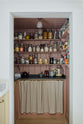 In a warm, inviting, muted yellow classic English kitchen features an walk-in, muted pink pantry with a single door base cabinet, an open base cabinet with an aged brass curtain rail and a black garnite counter, and three top shelves. Adorned with dried ingredients in a glass jards, a muted yellow tea set, a black ceramic tea pot, this kitchen feels humble and inviting for a family that enjoys cooking.