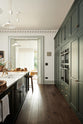 Looking between a large full-height pantry cupboard, oven cabinets, fridge/freezer cupboard, and a long kitchen island with a double sink, an aged brass Ionian tap in the grand bakehouse green classic English kitchen. Adorned with loaves of bread, leeks, potted plants, and still life paintings; the room feels homey and welcoming despite its large and decorative appearance.
