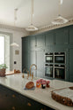 An oblique view of a long kitchen island with a double sink, an aged brass Ionian tap, the large full-height pantry cupboards, oven cabinets and fridge/freezer cupboards at the back of the grand bakehouse green classic English kitchen. Adorned with loaves of bread, a potted plant, still life paintings, and homemade jams, the room feels homey and welcoming despite its large and decorative appearance.