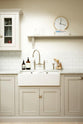 A close-up of a simple and stylish classic English kitchen’s double Belfast sink with antique silver Ionian tap, Carrara marble worktop, glazed wall cupboard, and shelf. Beautifully decorated with light copper rimmed clock, glass containers, wooden cleaning equipments, and white splashback tiles, it creates a functional and beautiful kitchen space.