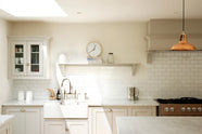 This close-up of a simple and stylish classic English kitchen highlights the sink and cooker run, featuring a double Belfast sink, an antique silver Ionian tap, and a Carrara marble worktop. It includes a glazed wall cupboard and a shelf. Beautifully decorated with a light copper pendant light, glass containers, white ceramics, a silver kettle, wooden cleaning equipment, and white splashback tiles, this space creates a functional and beautiful kitchen atmosphere.