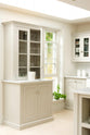 A close-up of a simple and stylish classic English kitchen’s glazed larder with a Carrara marble worktop. Beautifully decorated with white ceramics, drinking glasses, a basil plant, and white splashback tiles, it creates a functional and beautiful kitchen space.