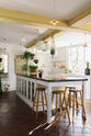 The white linen, classic English kitchen features an L-shaped sink and cooker run with Carrara marble worktop, a large single Carrara marble sink, an aged brass Ionian tap, and white crackle metro splashback tiles. In the centre is a large kitchen island with a solid oak worktop, breakfast nook and Helen stools. Lush green hanging plants and white porcelain lights, with the original yellow beam, enhance the bright and cheerful atmosphere, making the kitchen lively and inviting.