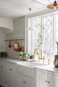 A close and angled look at a white linen, classic English kitchen features an L-shaped sink and cooker run with Carrara marble worktop, a large single Carrara marble sink, an aged brass Ionian tap, and white crackle metro splashback tiles. Lush green potted plants, white porcelain lights, and aged brass hanging rails with copper pots and pans enhance the bright and cheerful atmosphere, making the kitchen lively and inviting.