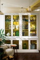 A large white linen curiosity cupboard with a yellow interior features hand-painted white flowers and green foliage, paying homage to the original yellow beams of the historical Millhouse building. It showcases beautiful vintage ceramics, glass centrepieces, framed detailed illustrations, and potted plants.