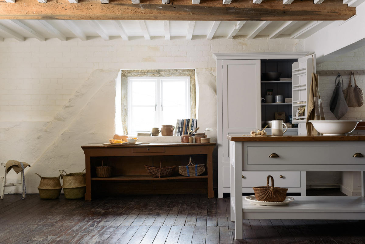 A beautiful white linen kitchen features a freestanding table, a pantry cupboard, and a collection of vintage furniture and items, reflecting on the well-reserved historical space with its original plastered walls, wooden beams, and oak floors.
